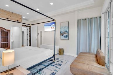 Sunlit modern coastal master bedroom with a black four-poster bed and crisp white bedding, leather bench by sliding glass doors, blue patterned rug, wall-mounted TV showing a beach scene, abstract ocean artwork, light blue curtains and wooden armoire.