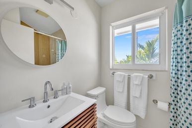 Bright modern coastal bathroom with white sink and wood vanity, round mirror, toilet, teal polka-dot shower curtain, and window view of palm trees and blue sky.