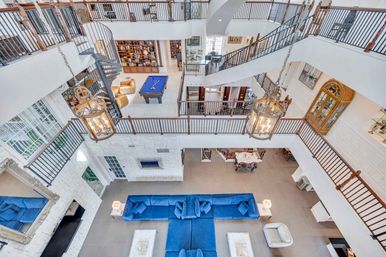 Aerial view of a luxury multi-level atrium living room featuring a large blue sectional sofa, hanging lantern chandeliers, a spiral staircase, upper gallery railings and a billiards table on the mezzanine