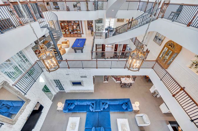 Aerial view of a luxury multi-level atrium living room featuring a large blue sectional sofa, hanging lantern chandeliers, a spiral staircase, upper gallery railings and a billiards table on the mezzanine