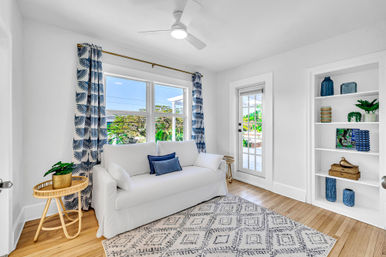Sunlit coastal-style living room with a white slipcovered sofa, blue patterned curtains, hardwood floors, geometric area rug, built-in shelves with blue decor, glass door to a patio, ceiling fan, and a round rattan side table with a potted plant.