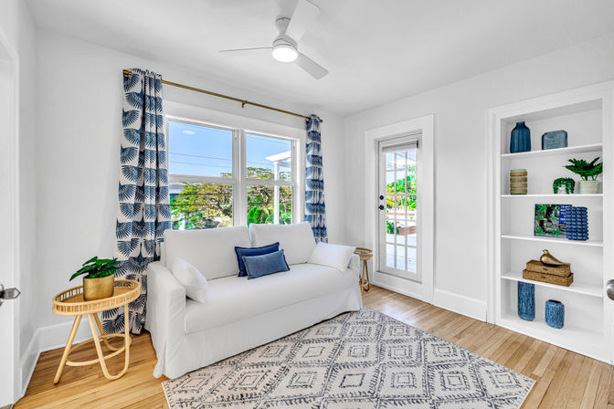 Sunlit coastal-style living room with a white slipcovered sofa, blue patterned curtains, hardwood floors, geometric area rug, built-in shelves with blue decor, glass door to a patio, ceiling fan, and a round rattan side table with a potted plant.