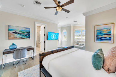 Bright beach-inspired master bedroom with king bed, wooden ceiling fan, wall-mounted TV, small desk with blue glass vase, ocean paintings, and a sunny windowed sitting nook with plantation shutters.