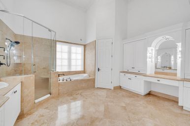 Bright luxury master bathroom with beige marble tile floors, glass-enclosed walk-in shower, built-in soaking tub under a frosted window, and white cabinetry vanity with arched mirror.