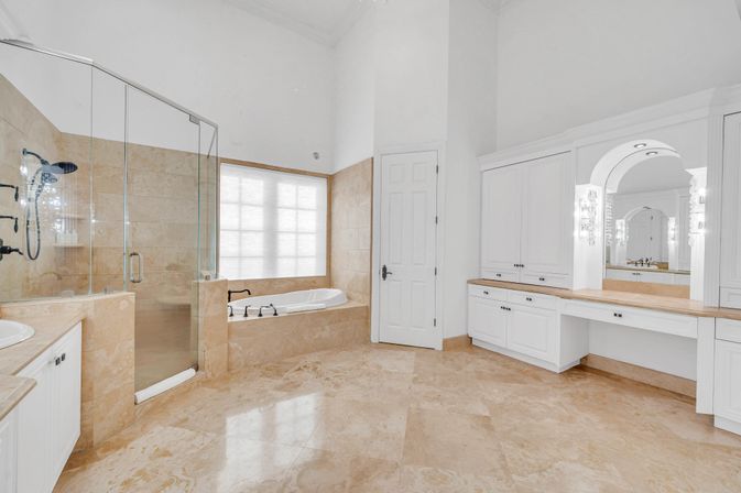 Bright luxury master bathroom with beige marble tile floors, glass-enclosed walk-in shower, built-in soaking tub under a frosted window, and white cabinetry vanity with arched mirror.