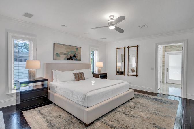 Bright contemporary master bedroom with upholstered platform king bed, dark hardwood floors, large area rug, twin nightstands with lamps, ceiling fan, abstract wall art and mirrors, doorway to en-suite bathroom.