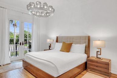 Sunlit modern bedroom with king bed and tufted headboard, crisp white linens and a mustard accent pillow, wooden nightstands with lamps, geometric chandelier, and French doors opening to a palm-lined yard.