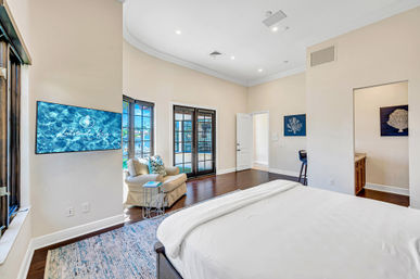 Sunlit coastal master bedroom with king bed, hardwood floors, cozy armchair, wall-mounted TV and French doors opening to a waterfront patio.