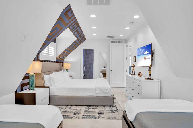 Bright coastal attic loft bedroom with sloped ceilings, king bed and two twin beds, geometric ceiling accent, white modern decor, bedside lamps and dresser with wall-mounted TV showing a beach scene.