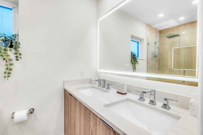 Bright modern bathroom with white double-sink vanity and wood cabinets, LED-framed mirror, chrome faucets, glass walk-in shower, small window with trailing plant, and wall-mounted toilet paper holder