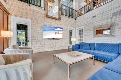 Bright coastal-chic living room with double-height light stone wall, blue velvet sectional, striped armchairs, marble-top coffee table, wall-mounted TV showing a beach scene, glass French doors and loft railing above.