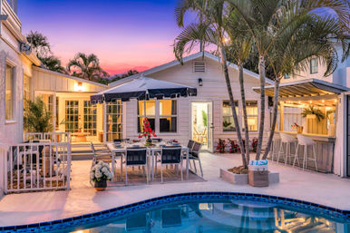 Tropical backyard patio at sunset with a curved swimming pool, outdoor dining table under a navy umbrella, bar with stools, palm trees and string lights.
