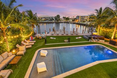 Sunset waterfront tropical backyard with illuminated palm trees, modern plunge pool, grassy lounge area with chairs and cornhole, and a wooden dock on a calm marina.