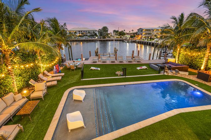 Sunset waterfront tropical backyard with illuminated palm trees, modern plunge pool, grassy lounge area with chairs and cornhole, and a wooden dock on a calm marina.