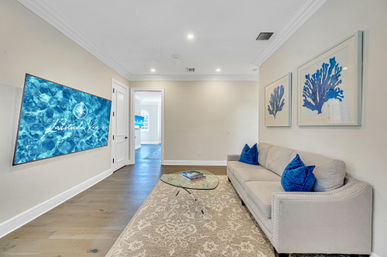 Coastal-style media room with a wall-mounted TV showing a blue water graphic, light gray sofa with bright blue pillows, glass coffee table on a patterned rug, hardwood floors and framed blue coral artwork on neutral walls.