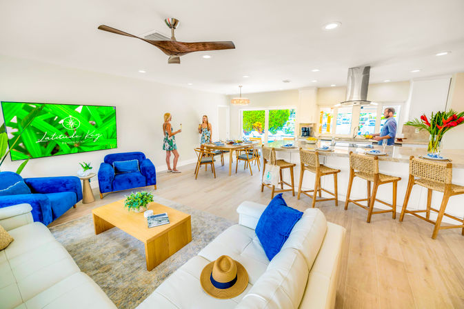 Sunny open-plan coastal vacation rental living room and kitchen with white leather sofas, bold blue accent chairs, wooden coffee table, breakfast bar with woven wicker stools, dining area, ceiling fan, and water view through sliding doors, three people casually socializing.