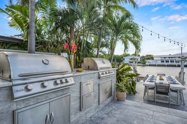 Waterfront tropical outdoor kitchen with stainless-steel double grills built into a stone counter, palm trees and potted plants, long dining table set on the dock under string lights by the water.