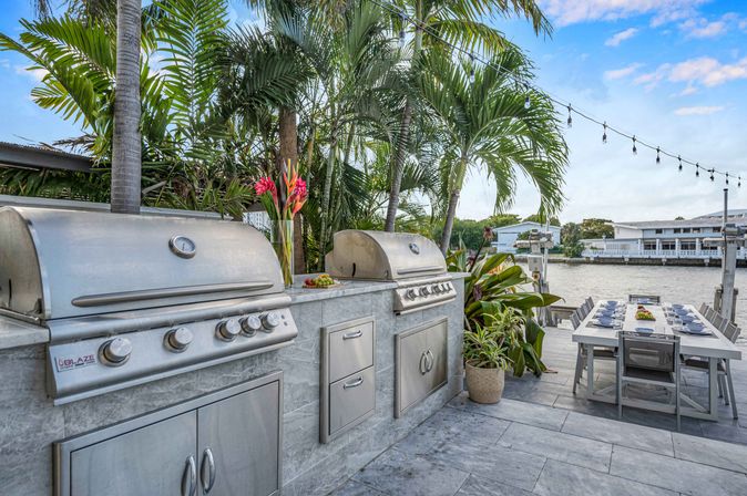 Waterfront tropical outdoor kitchen with stainless-steel double grills built into a stone counter, palm trees and potted plants, long dining table set on the dock under string lights by the water.