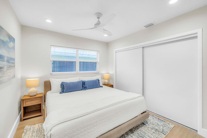 Bright modern coastal-style bedroom with white linens, blue accent pillows, wooden nightstands, table lamps, ceiling fan, and sliding closet doors