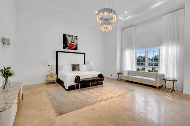 Bright, airy luxury master bedroom with high ceilings and crystal chandelier, tufted white king bed, velvet bench, cream rug, marble floors and a large window with a waterfront view