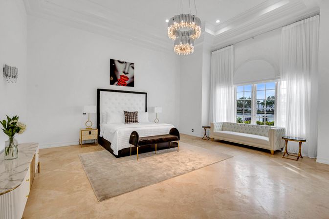 Bright, airy luxury master bedroom with high ceilings and crystal chandelier, tufted white king bed, velvet bench, cream rug, marble floors and a large window with a waterfront view