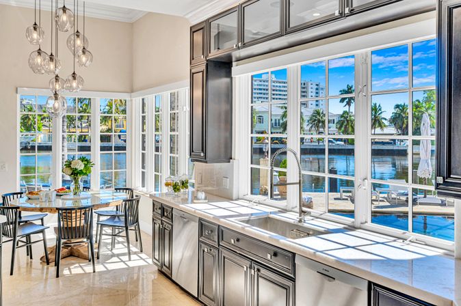 Sunlit coastal waterfront kitchen with marble countertops, stainless sink, dark cabinets and a round dining nook, large windows framing palm-lined canal and dock views.