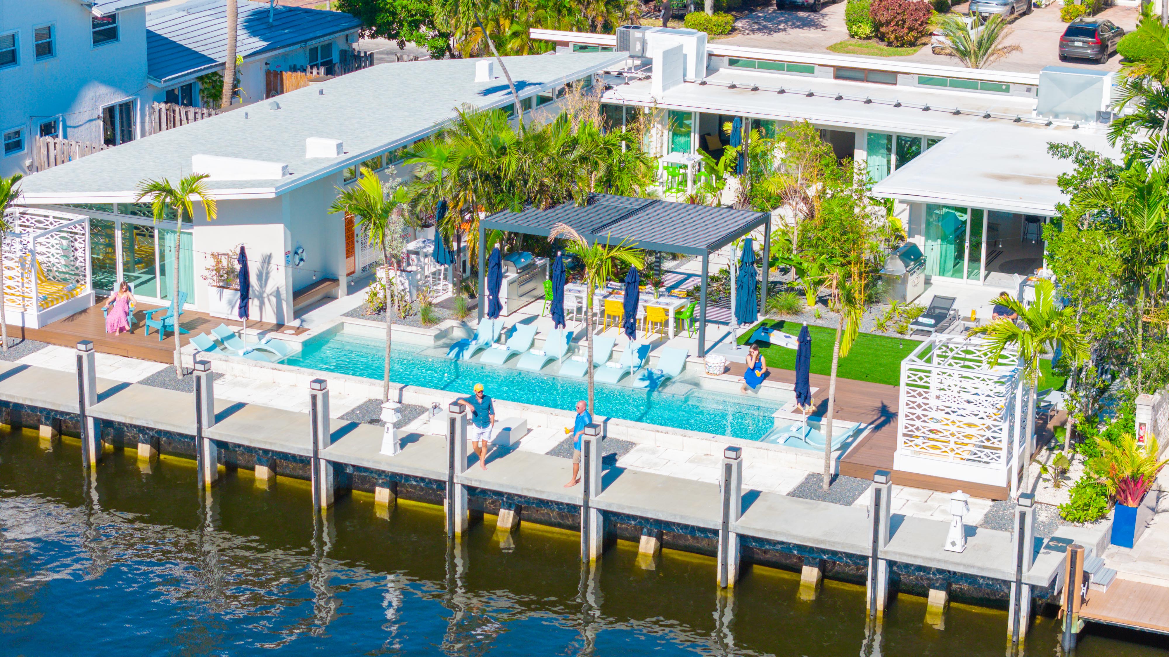 Sunny aerial view of a modern waterfront home with a turquoise rectangular pool, sun loungers under a pergola, palm trees, and a private dock along a calm canal