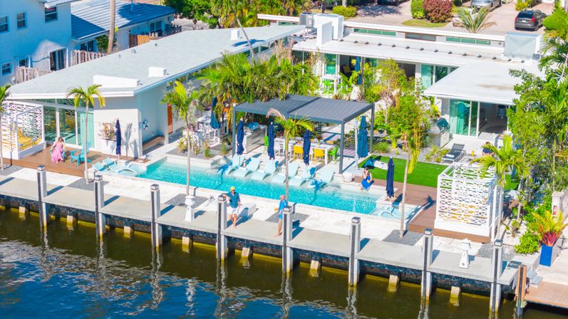 Sunny aerial view of a modern waterfront home with a turquoise rectangular pool, sun loungers under a pergola, palm trees, and a private dock along a calm canal