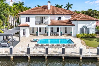 Sun-drenched Mediterranean-style waterfront villa with red tile roof, swimming pool and spa, row of lounge chairs, covered patio seating, private dock and palm trees