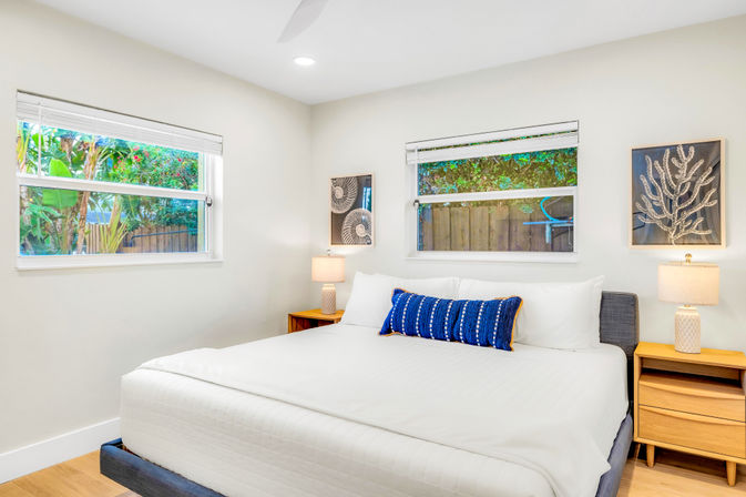 Sunny modern bedroom with king bed in crisp white linens, blue patterned lumbar pillow, wooden nightstands with lamps, coastal-style wall art, and two windows overlooking lush backyard greenery.
