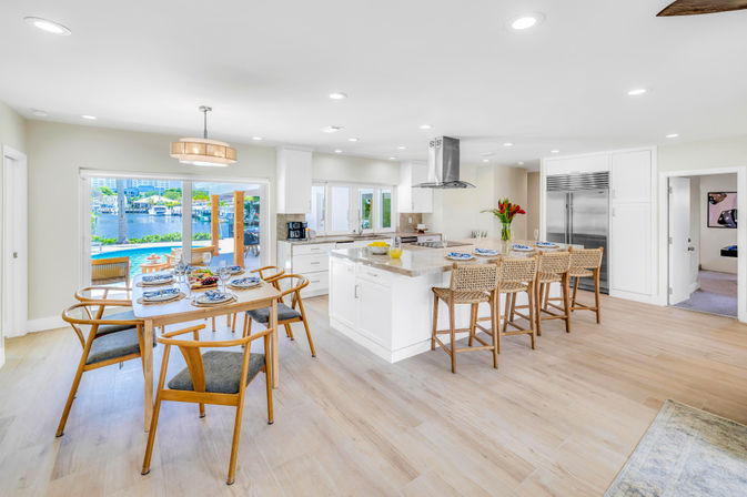 Bright modern open-concept waterfront kitchen and dining area with white island and stone countertops, woven bar stools, wooden dining table set, and sliding glass doors opening to a pool and marina view