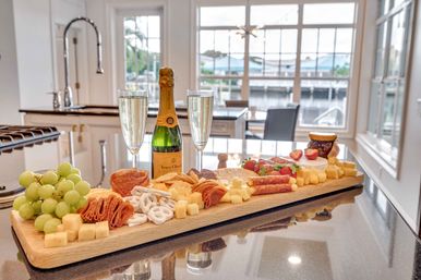 Gourmet charcuterie board on a glossy kitchen island with green grapes, cubed cheeses, sliced salami, pretzels, strawberries and jam, paired with a bottle of champagne and two flutes in a bright waterfront kitchen with large windows.