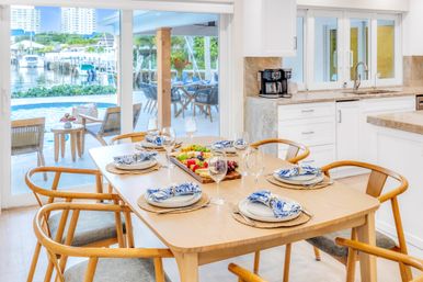 Sunlit coastal dining room with a wooden table set for six—blue-pattern napkins, wine glasses and a fruit platter—sliding glass doors opening to a poolside patio and waterfront marina view.