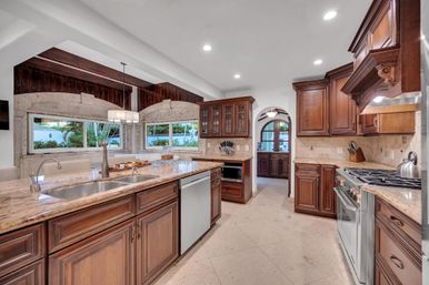 Chef-ready luxury kitchen with long marble island and undermount sink, rich wood cabinetry, stainless steel range and dishwasher, arched doorway, tile floors and wide windows overlooking lush landscaping