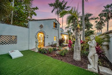 Sunset Mediterranean-style home with ornate arched iron gate, tropical palms, manicured turf and classical stone statues in a landscaped garden.