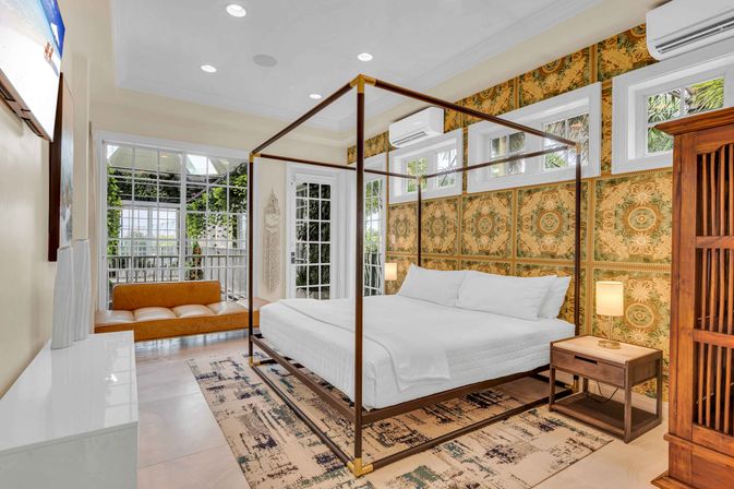 Sunlit tropical-inspired bedroom with a metal four-poster bed, crisp white linens, patterned golden wallpaper, wooden nightstand and leather bench by glass doors opening to a lush patio — coastal retreat