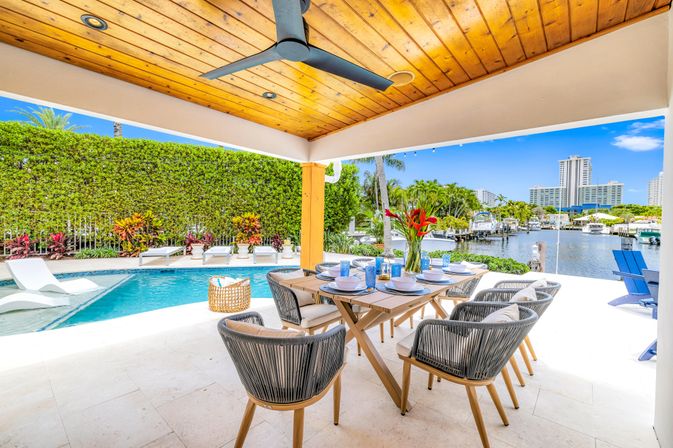 Sunny luxury waterfront patio with wooden ceiling and fan, outdoor dining table set for eight, adjacent pool with in‑water loungers, lush tropical hedges and canal with boats and distant high‑rises.