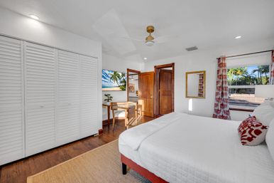 Sunlit coastal bedroom with white quilted bed, red patterned pillows and curtains, hardwood floors, white louvered closet, wood desk and chair, ceiling fan, wall-mounted TV showing a beach, and a window view of palm trees.