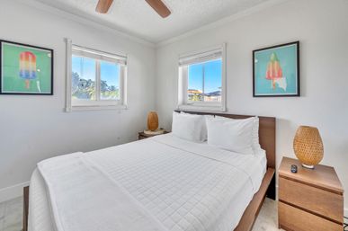 Bright beachside vacation rental bedroom with queen bed in crisp white linens, wood headboard and nightstands, woven rattan lamps, colorful popsicle artwork, and two windows framing palm trees and blue sky.