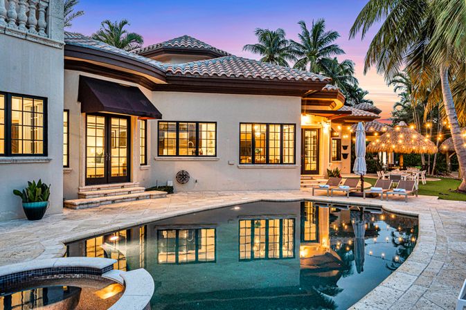 Luxury Mediterranean-style coastal backyard oasis at sunset — tiled-roof villa with warm lit windows reflected in a curved swimming pool and spa, poolside loungers, tiki-hut dining area, palm trees and string lights.