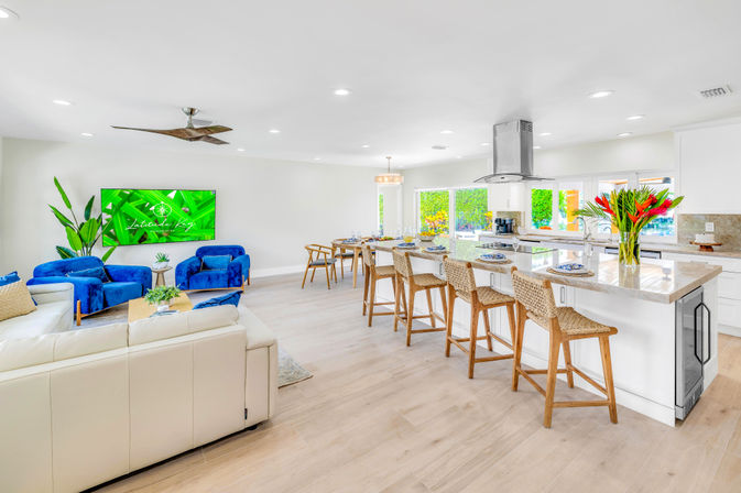 Sunlit open-plan living room and kitchen with light wood floors, white leather sofa, two royal-blue velvet armchairs, large marble island with woven barstools, stainless range hood, wall-mounted TV, tropical plants and red flower arrangement.