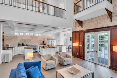 Airy open-plan waterfront living room and gourmet kitchen with double-height ceiling and balcony — blue velvet sofa, two swivel armchairs, marble coffee table, white island with stainless appliances and brick backsplash, and French doors opening to a waterside patio.