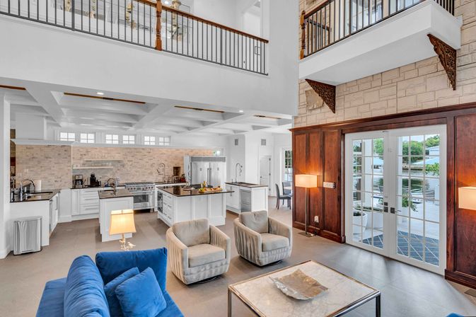 Airy open-plan waterfront living room and gourmet kitchen with double-height ceiling and balcony — blue velvet sofa, two swivel armchairs, marble coffee table, white island with stainless appliances and brick backsplash, and French doors opening to a waterside patio.