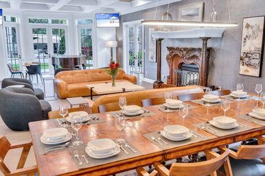 Sunlit open-plan dining and living room: long wooden table set for eight, caramel leather sofas, plush gray lounge chairs, ornate carved fireplace, grand piano, and floor-to-ceiling windows overlooking a garden.