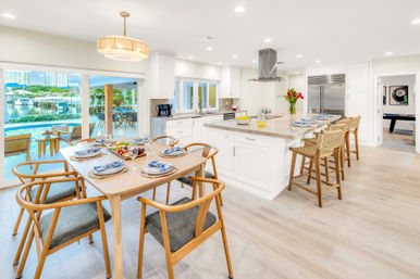 Sunny open-plan coastal kitchen and dining area with white island and wicker bar stools, wooden table set for six, sliding glass doors opening to a pool and marina waterfront view with boats and condos.
