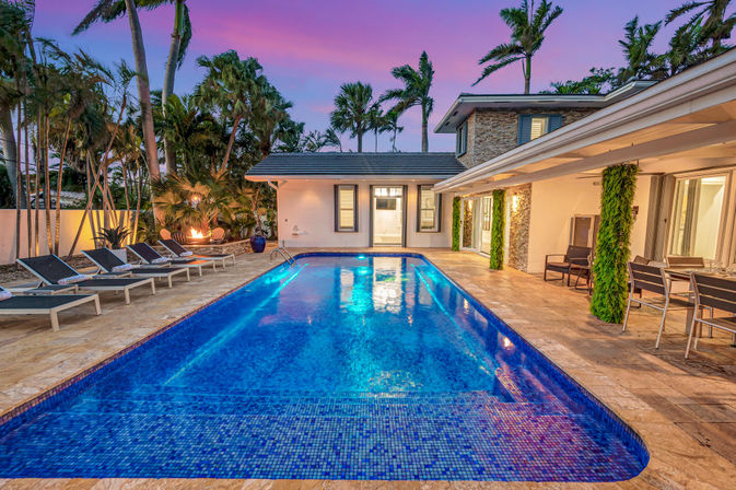 Luxury tropical backyard oasis at dusk with a rectangular blue-tiled swimming pool, poolside loungers, covered patio dining, palm trees and a purple-pink sunset sky.
