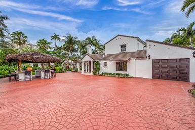 White stucco Mediterranean-style home in a tropical South Florida setting with a wide red stamped-concrete driveway, a thatched tiki bar with stools, palm trees and blue sky.