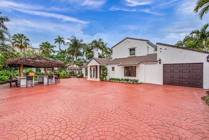 White stucco Mediterranean-style home in a tropical South Florida setting with a wide red stamped-concrete driveway, a thatched tiki bar with stools, palm trees and blue sky.