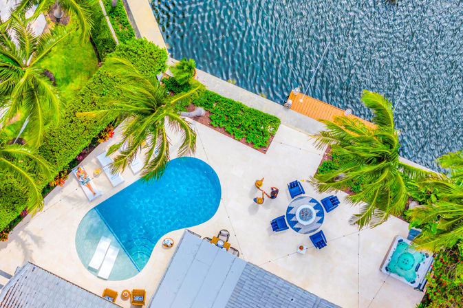 Aerial view of a vibrant tropical waterfront patio with a kidney-shaped pool, swaying palm trees, lounge chairs, a round blue seating area with fire pit, hot tub, and a small wooden dock on rippling water.