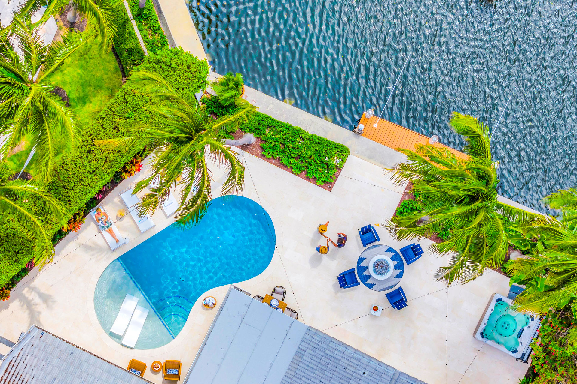 Aerial view of a vibrant tropical waterfront patio with a kidney-shaped pool, swaying palm trees, lounge chairs, a round blue seating area with fire pit, hot tub, and a small wooden dock on rippling water.